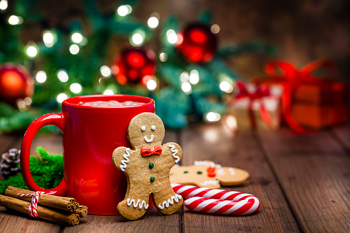 Christmas backgrounds: gingerbread cookies and hot chocolate shot on rustic wooden table. The composition is at the left of an horizontal frame leaving useful copy space for text and/or logo at the right. Christmas tree and string lights are out of focus at background.  Predominant colors are red, green and brown.